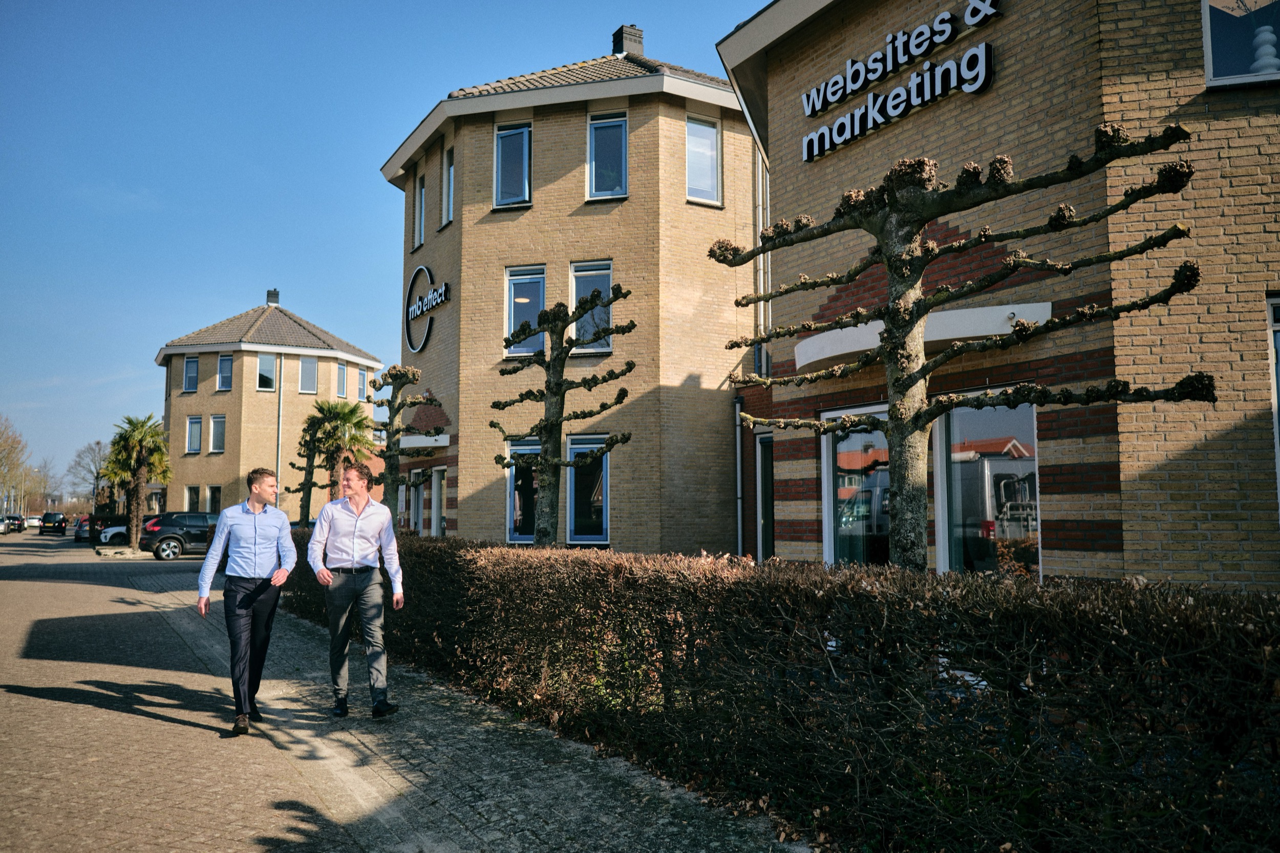 Twee professionele mannen lopen samen voor modern kantoorpand met "websites & marketing" signage, Leerdam — Stefan Segers Fotografie