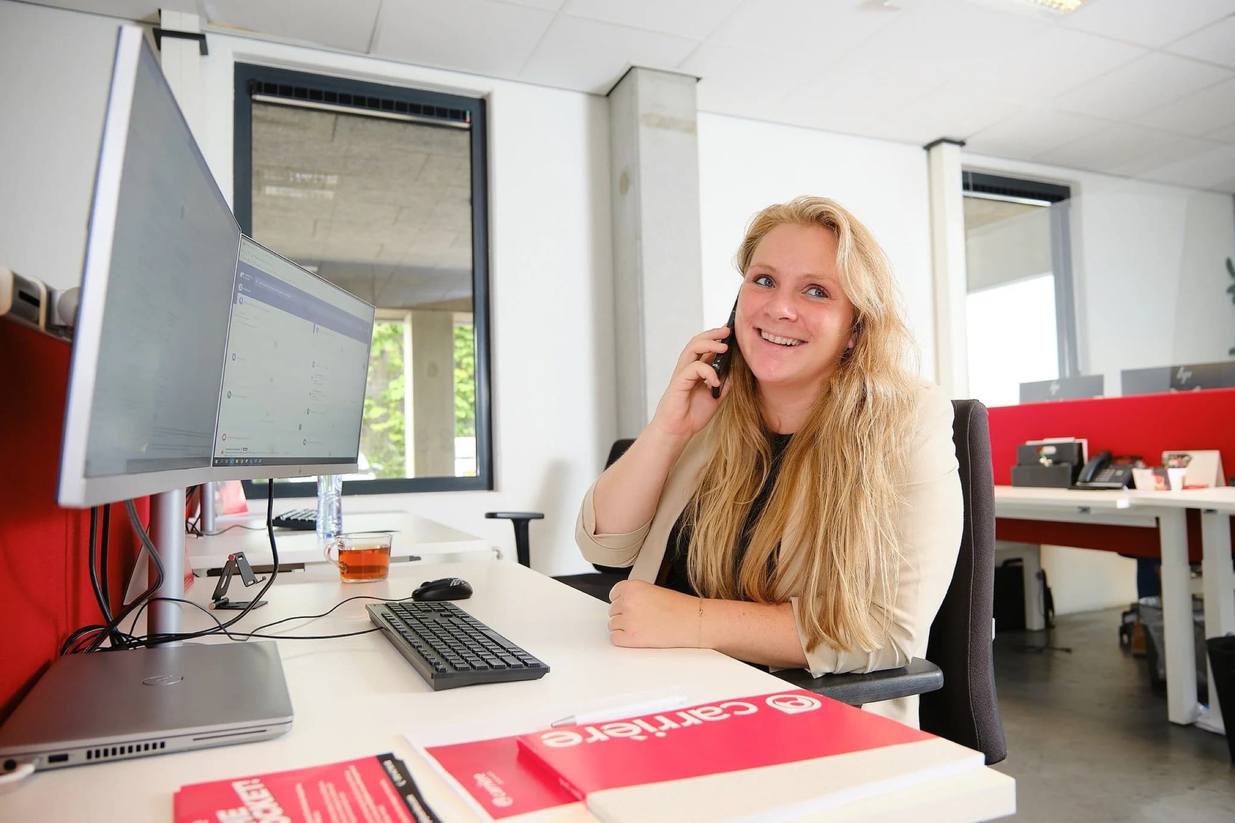 Vrouw aan bureau met monitor glimlacht naar camera, moderne kantooromgeving met rode accenten — Stefan Segers Fotografie