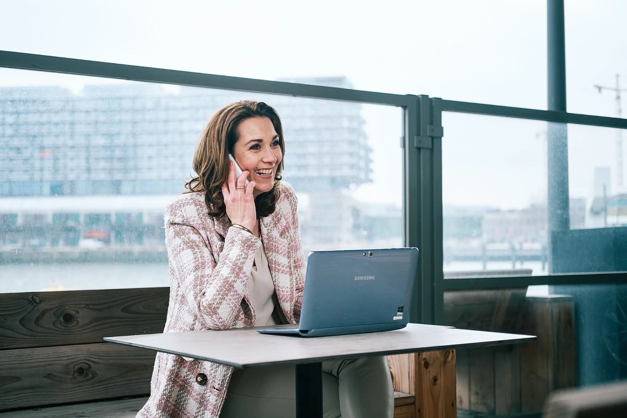Vrouw in roze blazer aan tafel met laptop, telefoonoproep, uitzicht op stadssilhouet — Stefan Segers Fotografie