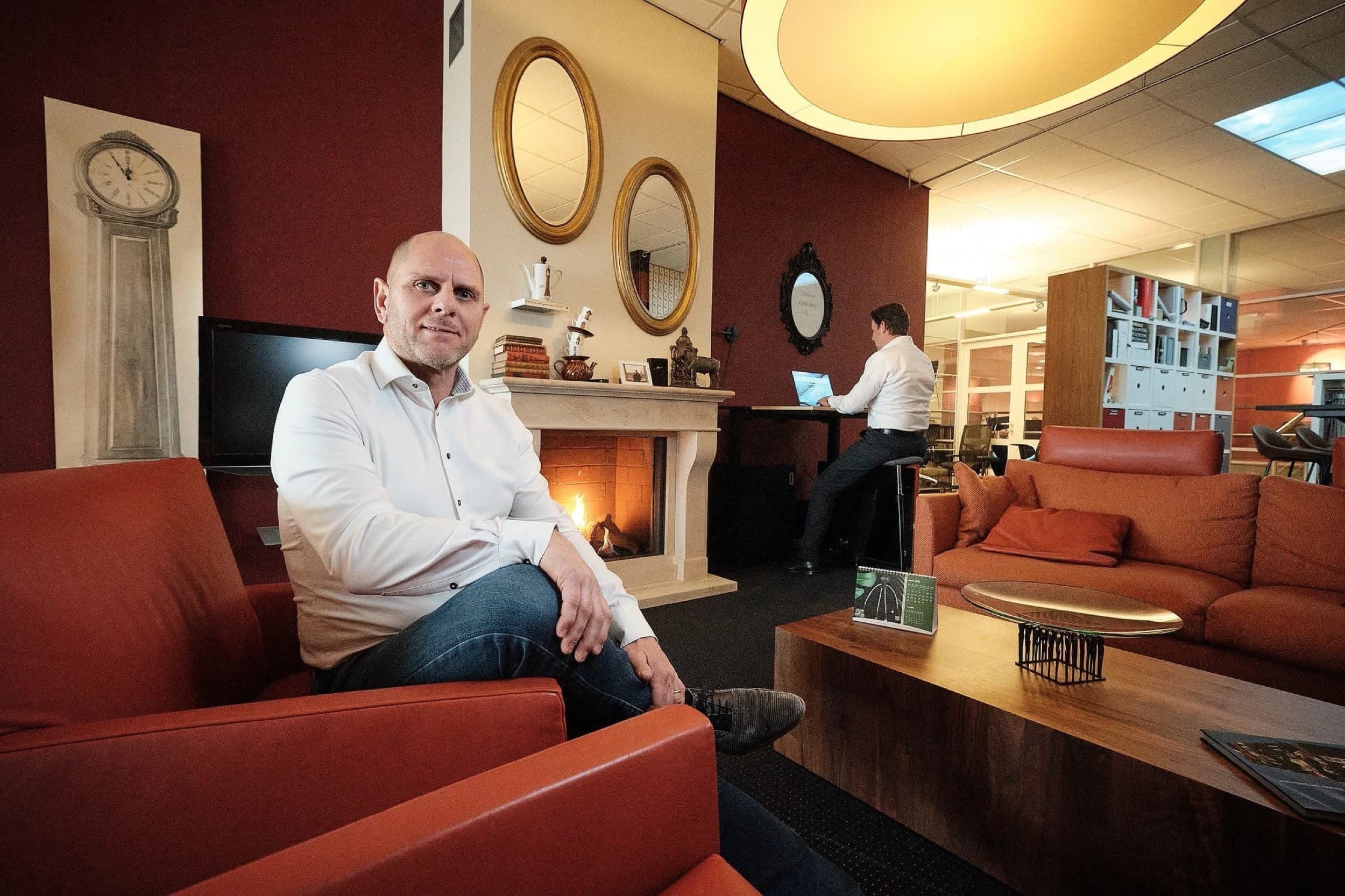 Man in wit overhemd zit ontspannen op oranje loungebank in modern kantoor met haard, collega op achtergrond — Stefan Segers Fotografie