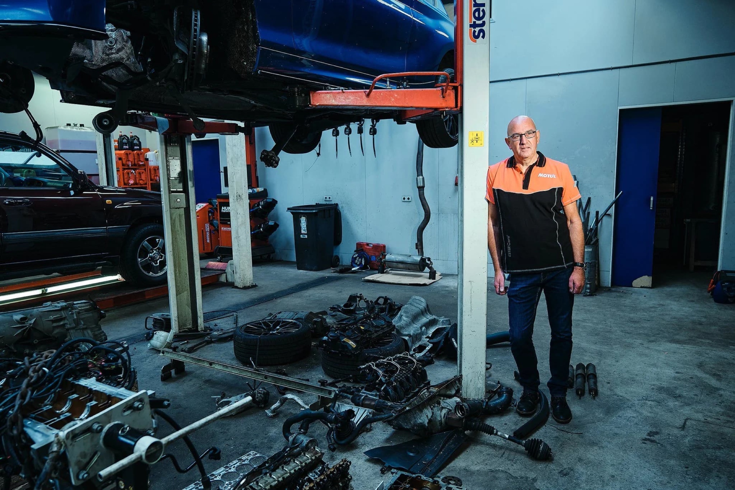 Monteur in oranje Motul werkkleding staat tussen auto-onderdelen in autowerkplaats — Stefan Segers Fotografie