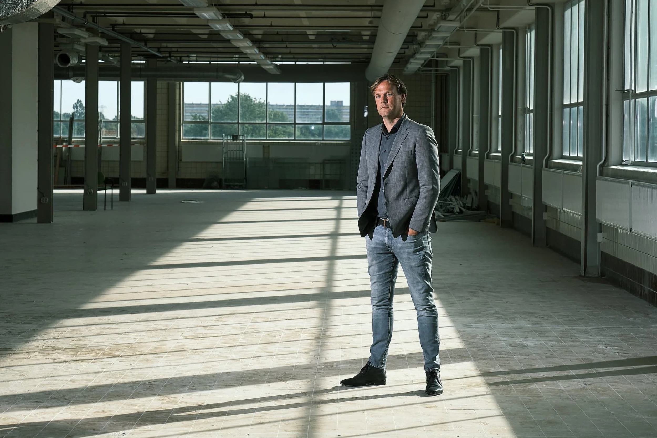 Man in grijs blazer staat zelfverzekerd in modern leeg kantoorruimte met grote ramen, bedrijfsreportage — Stefan Segers Fotografie