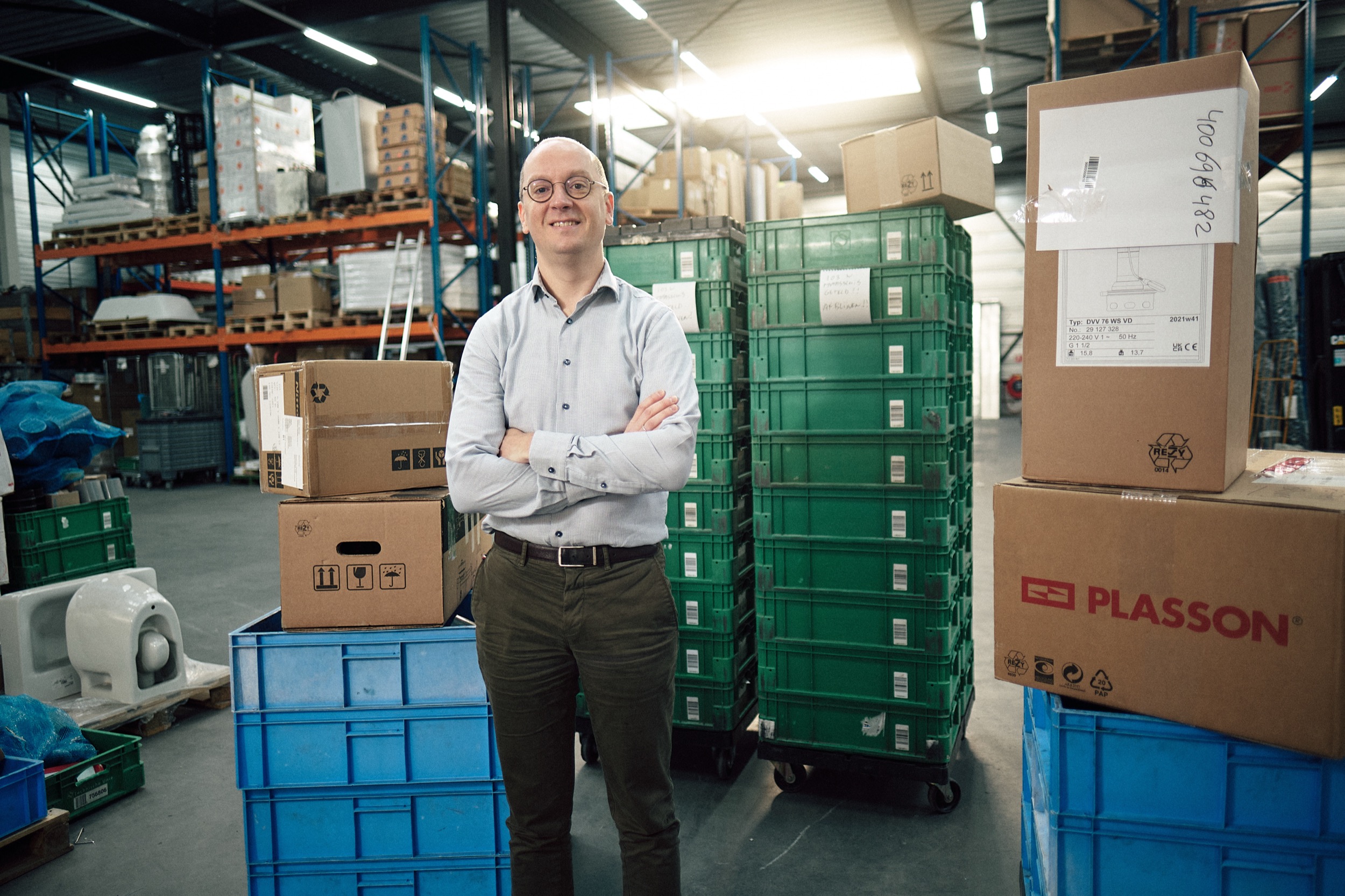 Man in licht overhemd staat zelfverzekerd in industrieel magazijn met kartonnen dozen en groene kratten, Rotterdam — Stefan Segers Fotografie