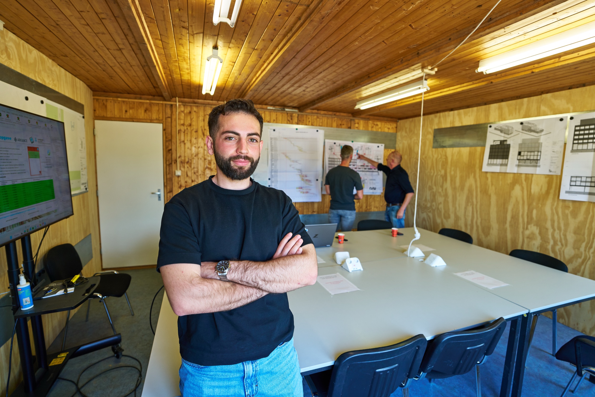 Man met gekruiste armen poseert in houten bouwkeet met collega's op achtergrond, Geleen — Stefan Segers Fotografie