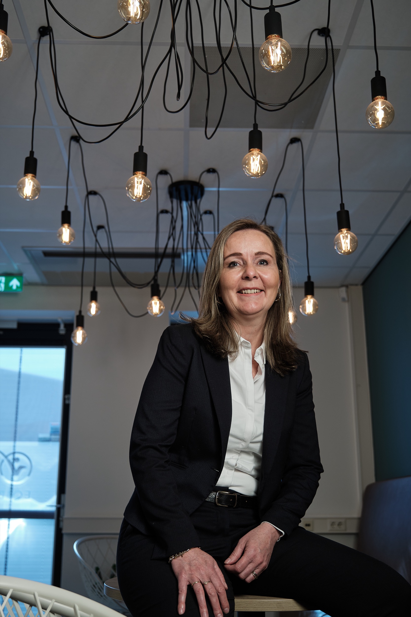 Professionele vrouw in donker pak en wit overhemd zit onder designverlichting, portretfoto voor Facto magazine — Stefan Segers Fotografie