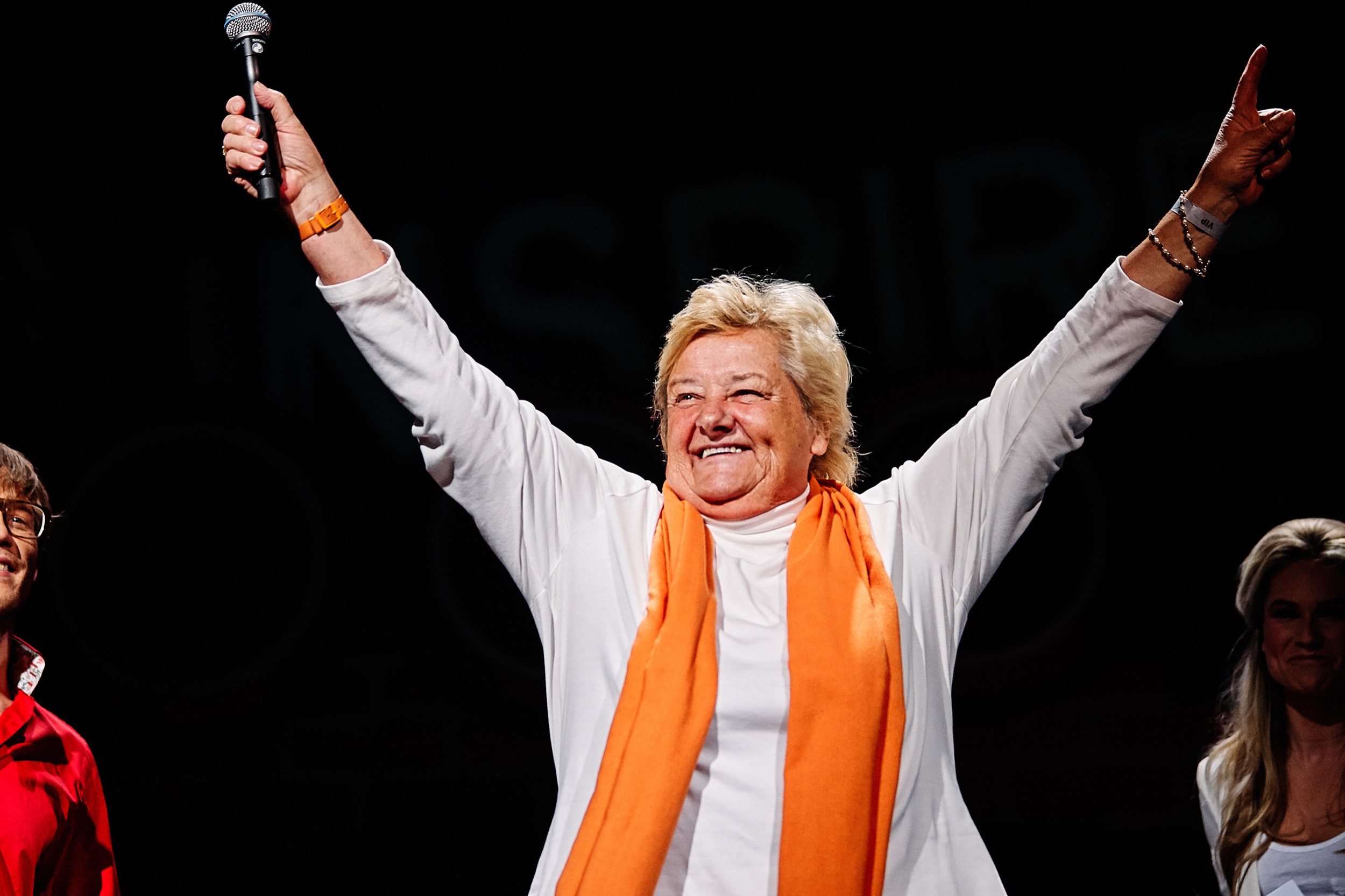 Vrouw in wit jasje en oranje sjaal heft armen triomfantelijk omhoog met microfoon op podium, evenement Dance4Life Ahoy Rotterdam — Stefan Segers Fotografie