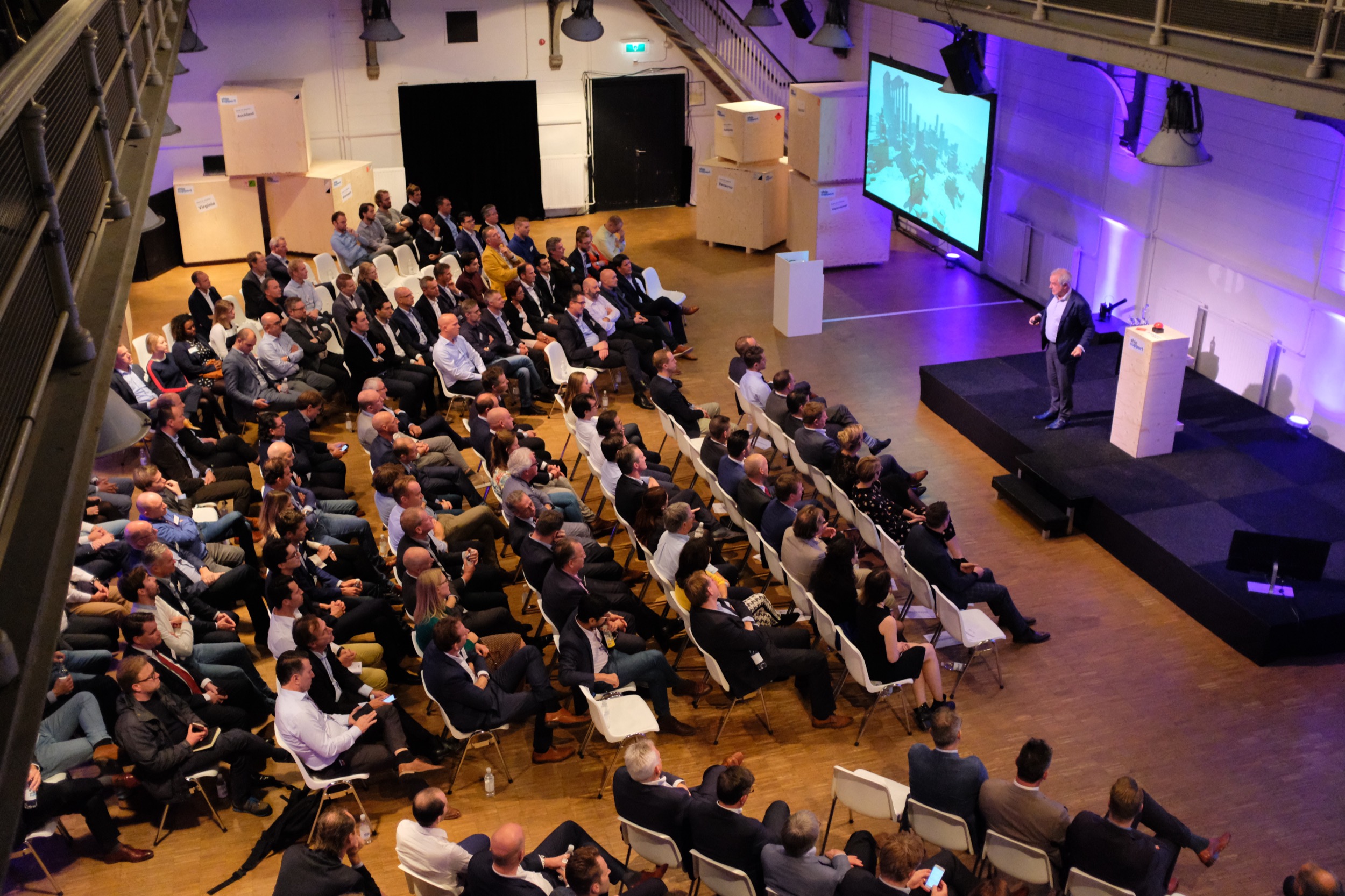 Spreker presenteert op podium voor groot zakelijk publiek in moderne zaal met paarse verlichting, evenementfotografie — Stefan Segers Fotografie