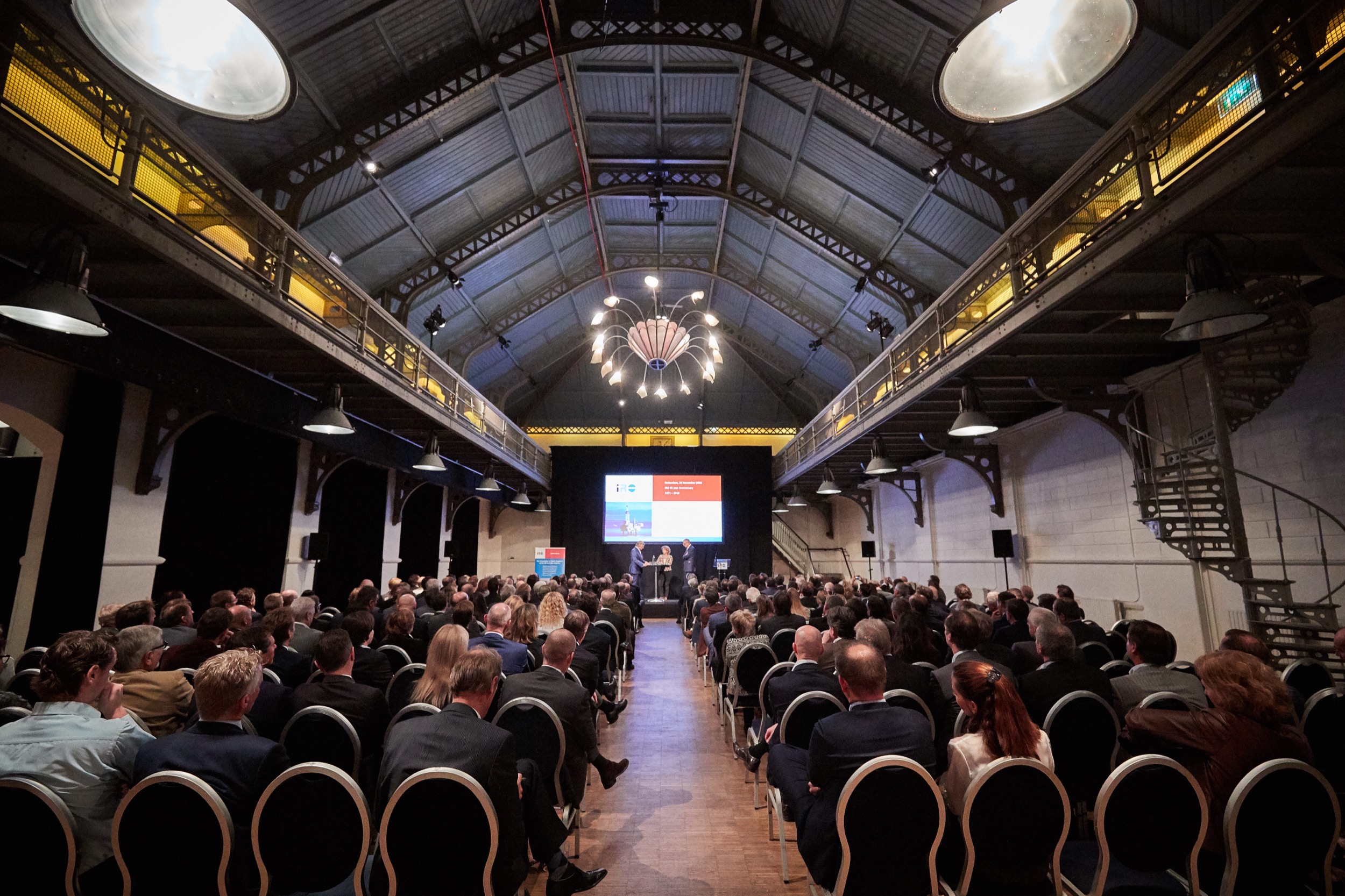Groot zakelijk evenement met honderden gasten in moderne hal, spreker op podium met scherm, industriële plafondarchitectuur Rotterdam — Stefan Segers Fotografie