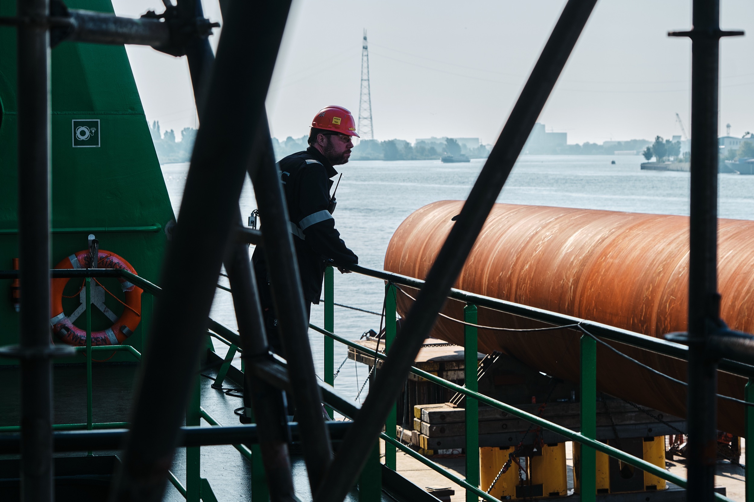 Arbeider in rode helm inspecteert grote koperen pijp op scheepswerf aan waterkant — Stefan Segers Fotografie