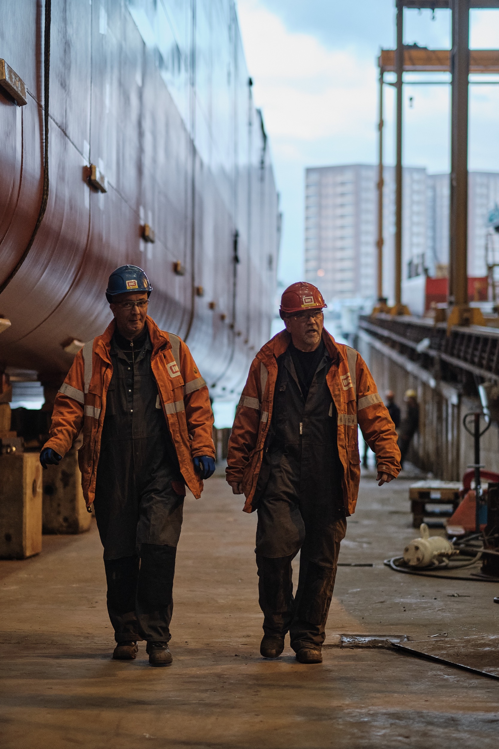 Twee werknemers in oranje veiligheidspak lopen langs industriële scheepsromp in fabriekshal Kinderdijk — Stefan Segers Fotografie