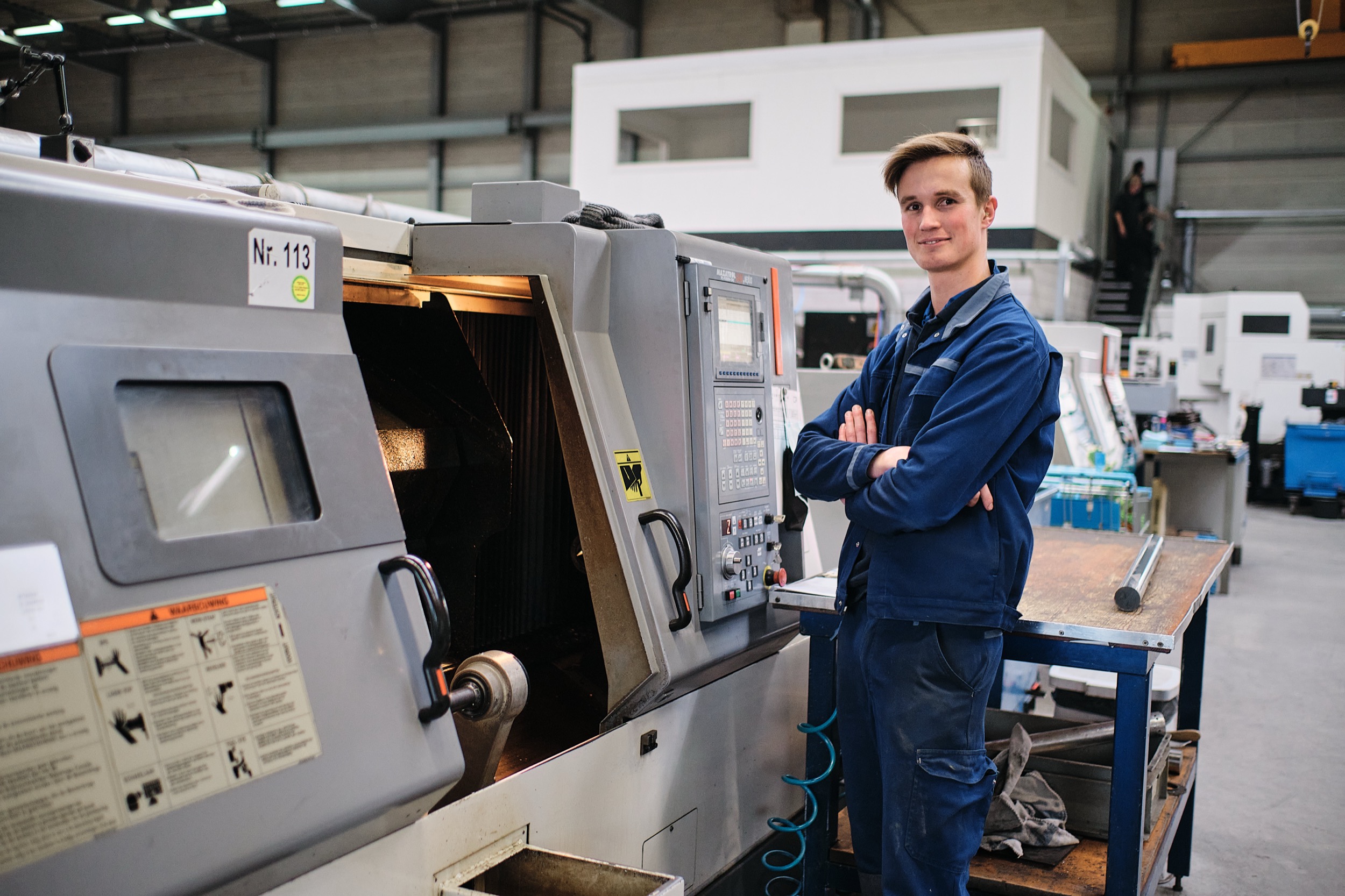 Jonge technicus in blauwe werkkleding staat zelfverzekerd naast CNC-machine in industriële fabriekshal, De Lier — Stefan Segers Fotografie