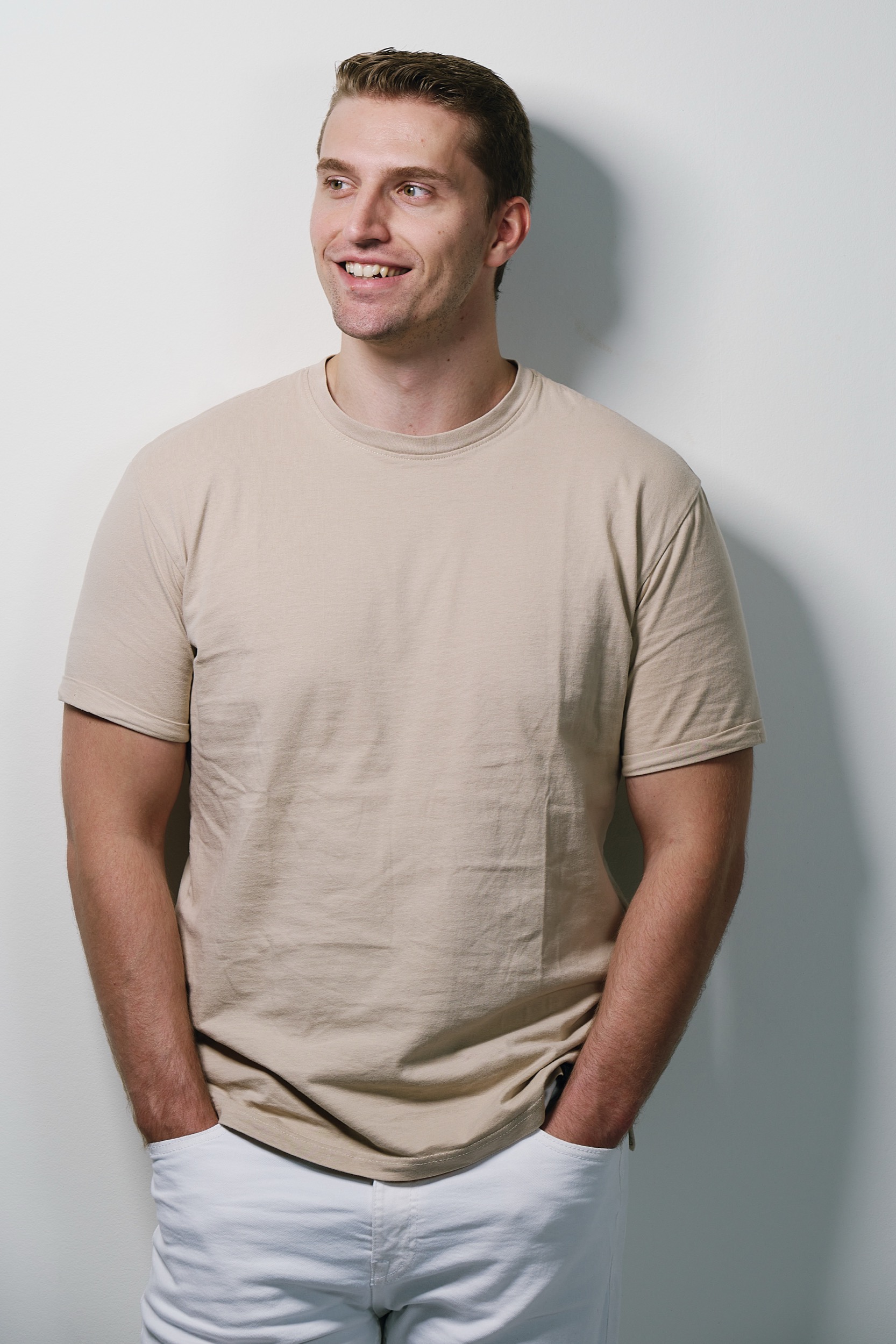 Man in beige t-shirt en witte broek glimlacht naar links, studioverlichting, persportret DJ Unphased — Stefan Segers Fotografie