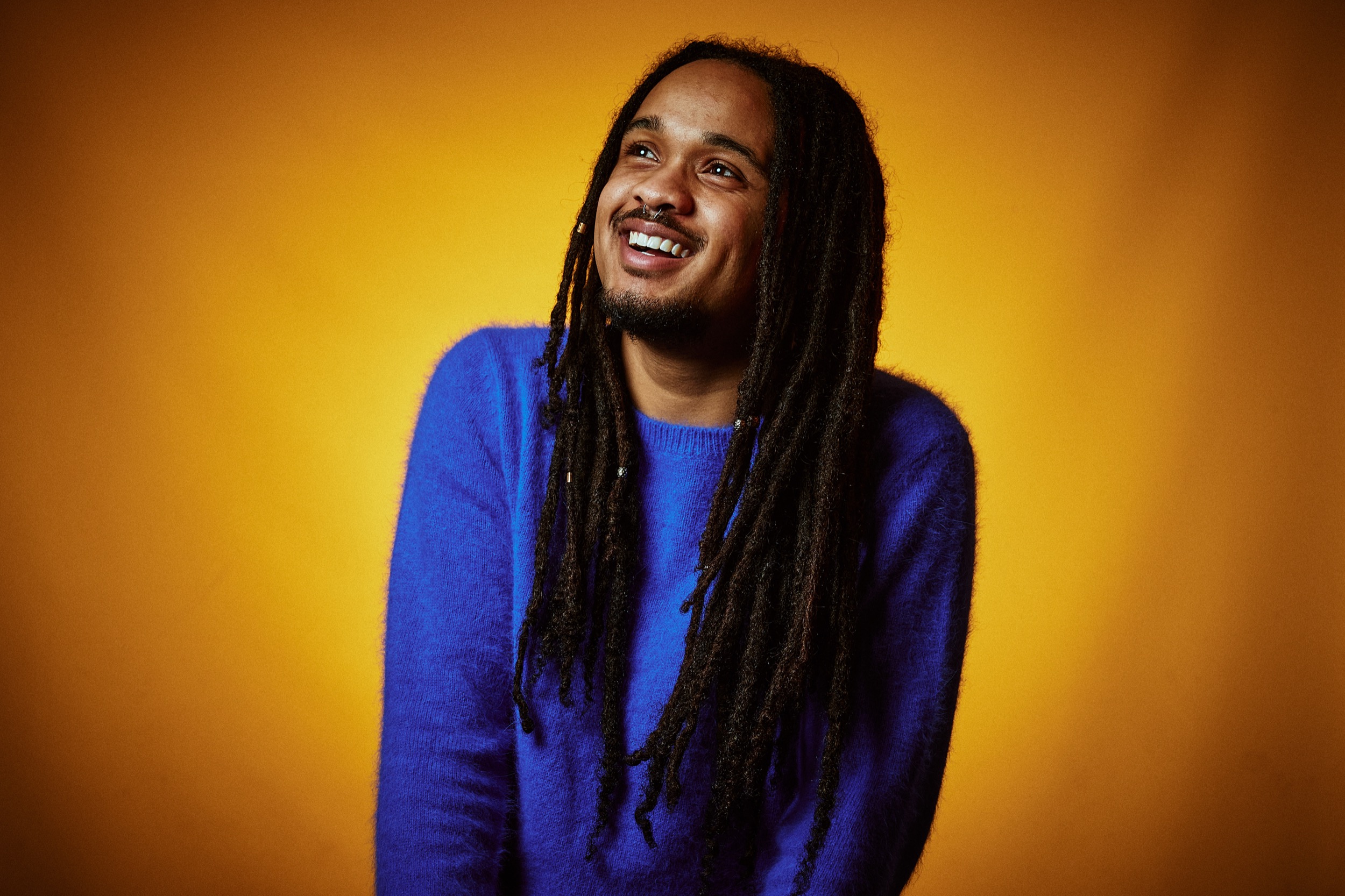 Man met dreadlocks in blauw shirt lacht tegen gele achtergrond, professioneel artistenportret, Zoetermeer — Stefan Segers Fotografie