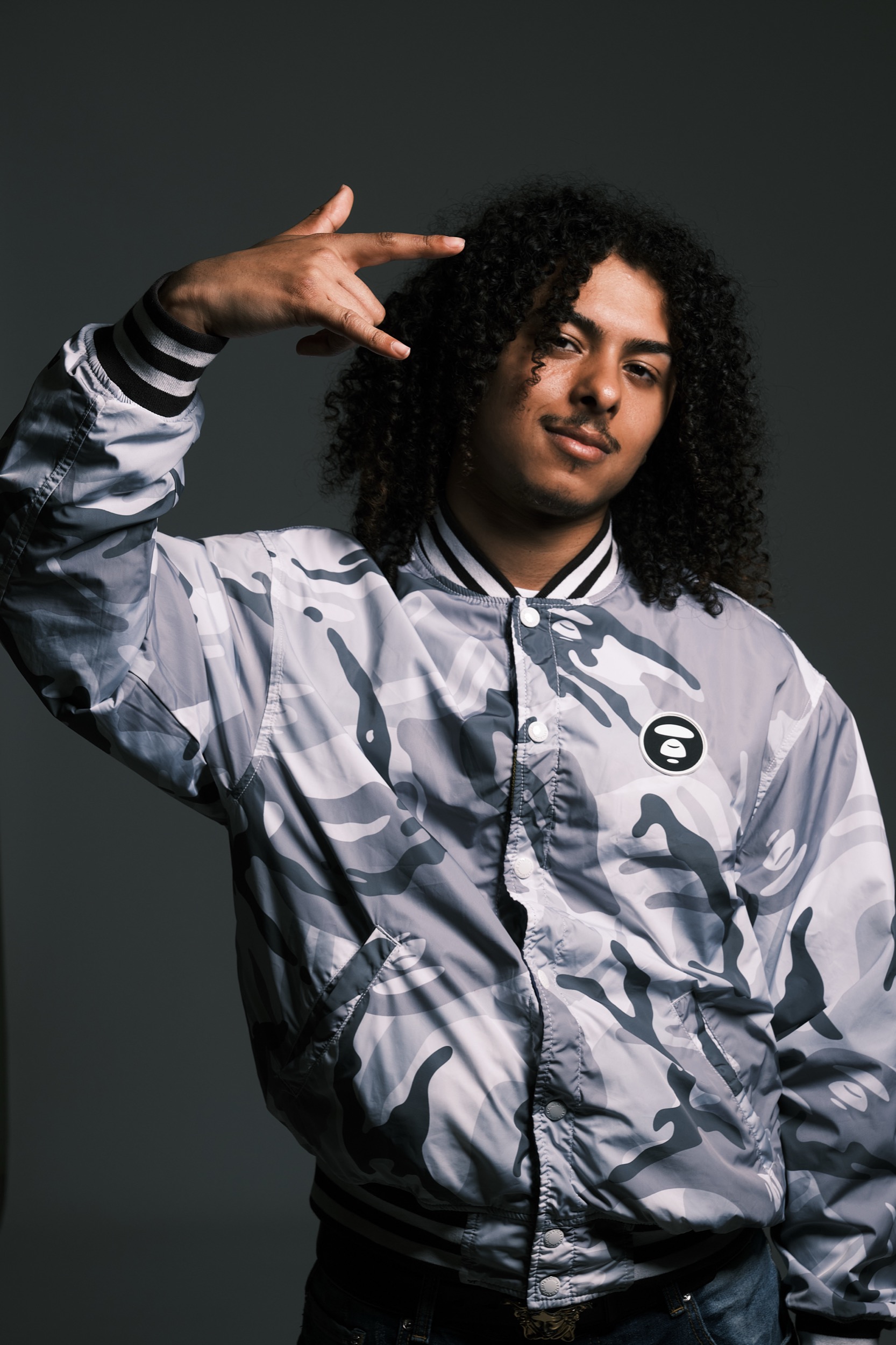 Jonge man met krullen in wit camouflage bomber jacket maakt peace-gebaar, studioportret — Stefan Segers Fotografie