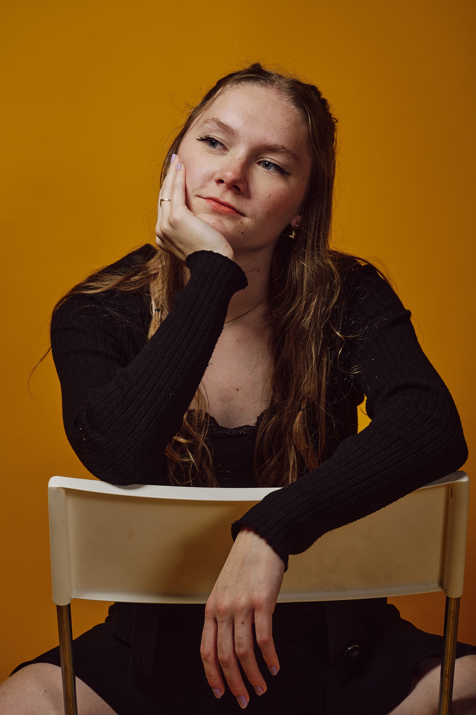 Vrouw zit op witte stoel tegen gele achtergrond, peinzende blik, zwarte kleding, artiest portret — Stefan Segers Fotografie