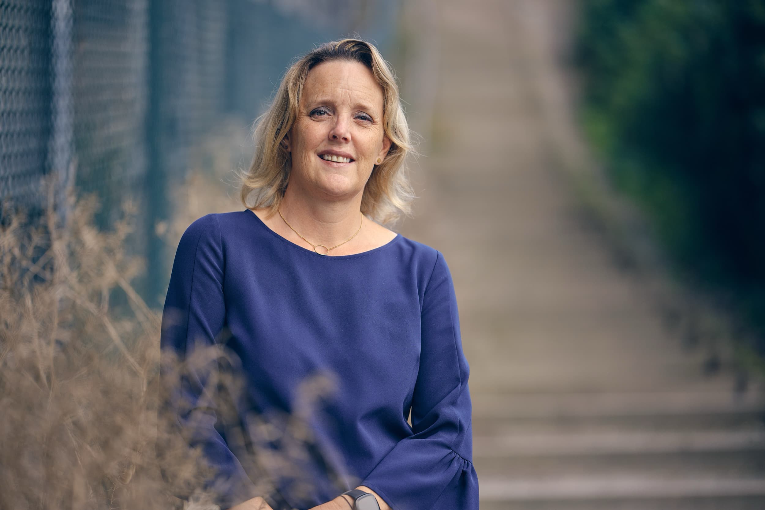Vrouw in donkerblauw shirt lacht naar camera buiten tegen natuurlijke achtergrond, professionele profielfoto — Stefan Segers Fotografie